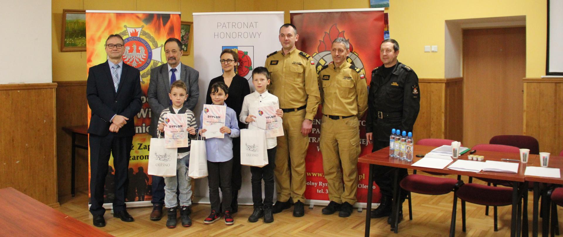 W sali w Miejskim Domu Kultury w Oleśnie są ustawieni w szeregu laureaci I kategorii wiekowej etapu gminnego OTWP wraz z opiekunem, Komendant Powiatowy Państwowej Straży Pożarnej w Oleśnie i jego zastępca, Komendant Gminny OSP Gminy Olesno, sekretarz jury OTWP oraz zastępca dowódcy Jednostki Ratowniczo-Gaśniczej w Oleśnie. W tle stoją 3 banery: OTWP, Burmistrza Olesna i Komendy Powiatowej Państwowej Straży Pożarnej w Oleśnie.