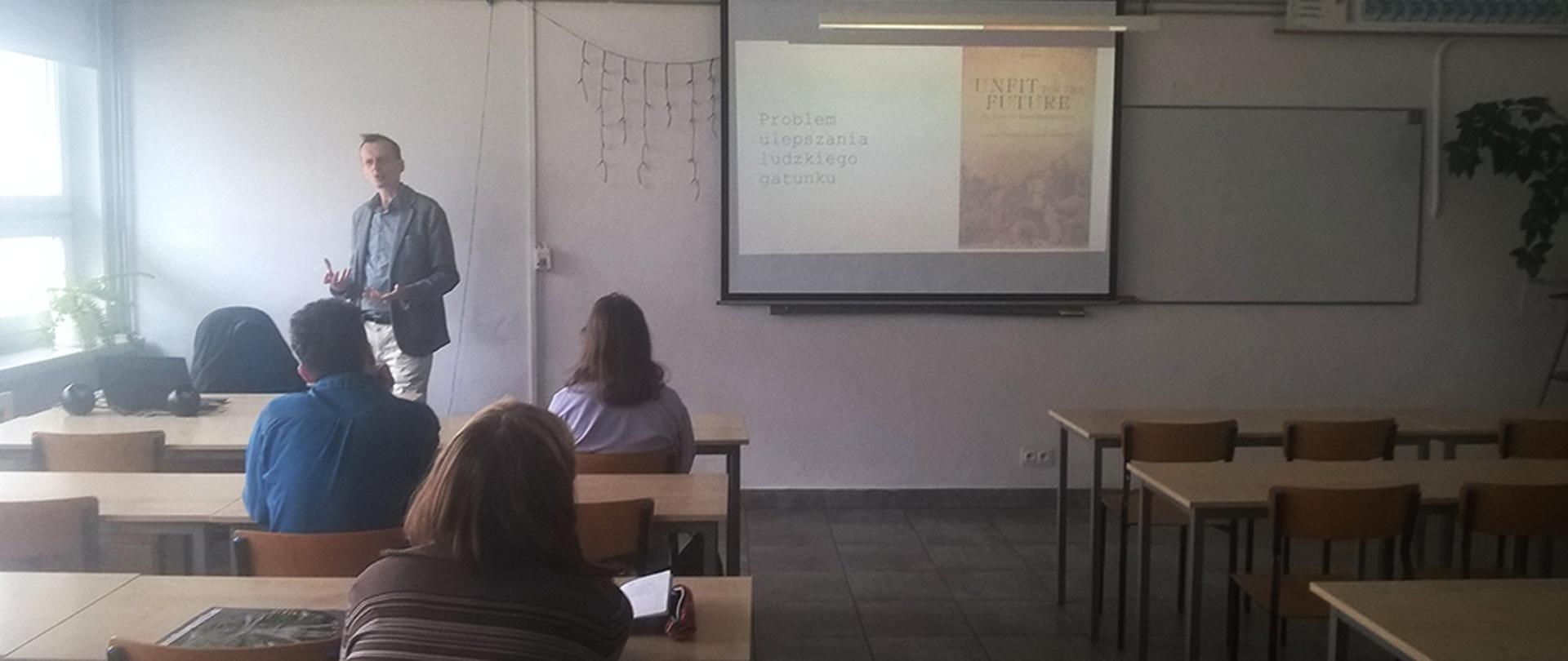 Wykładowca stojący przed grupą studentów w sali wykładowej, prowadzący prezentację multimedialną na temat „Problem ulepszania ludzkiego gatunku”.