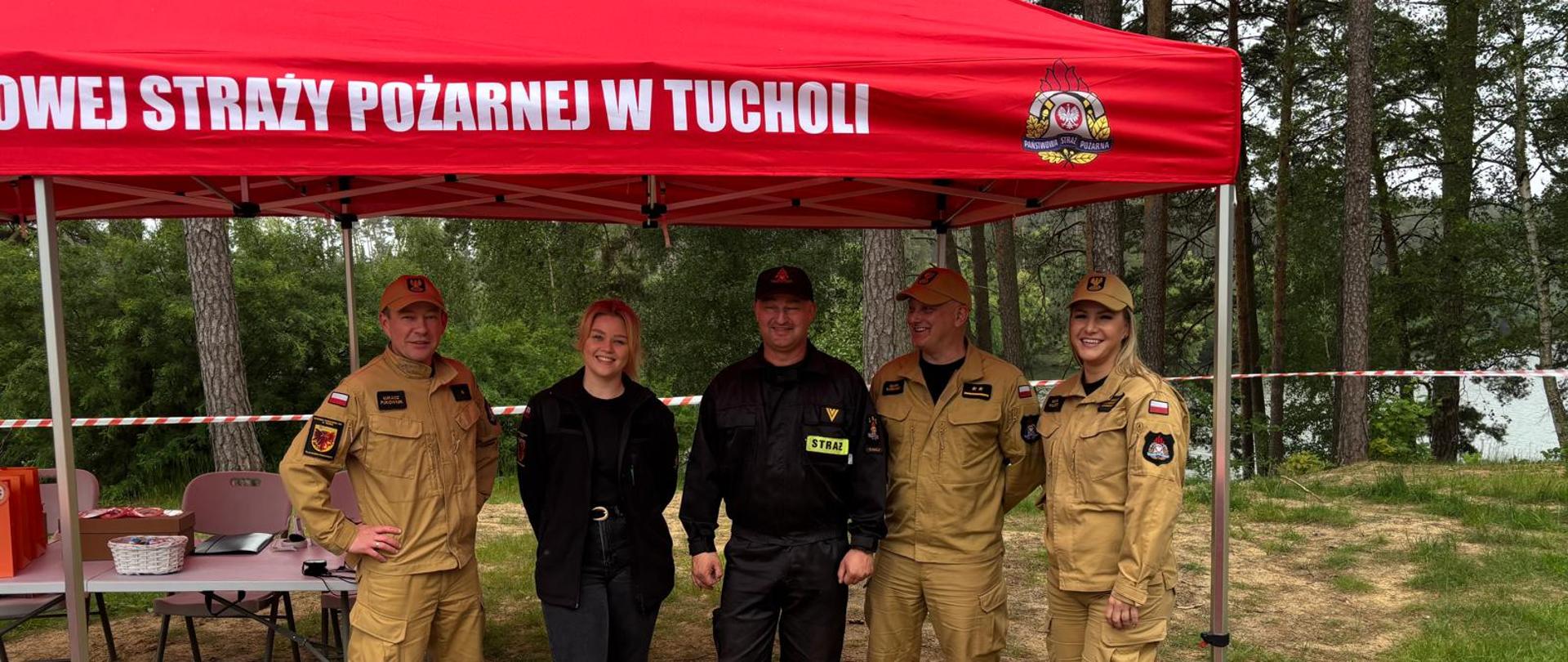 Pięć osób stoi pod czerwonym namiotem. Z lewej strony stoi strażak w mundurze koloru piaskowego. Obok stoi kobieta ubrana na czarno. Obok kobiety z prawej strony stoi strażak ubrany w umundurowanie koloru czarnego. Następnie z prawej strony stoi strażak i kobieta strażak ubrani w umundurowanie koloru piaskowego. Na namiocie widnieje napis ,,Komenda Powiatowa Państwowej Straży Pożarnej w Tucholi” oraz z prawe strony logo Państwowej Straży Pożarnej. Z lewej strony za grupą ludzi widać szary stolik. Za stolikiem widać dwa szare krzesła. Na stoliku widać postawiony biały koszyk, brązowy karton oraz pomarańczowe torebki prezentowe. W tle widać drzewa. 