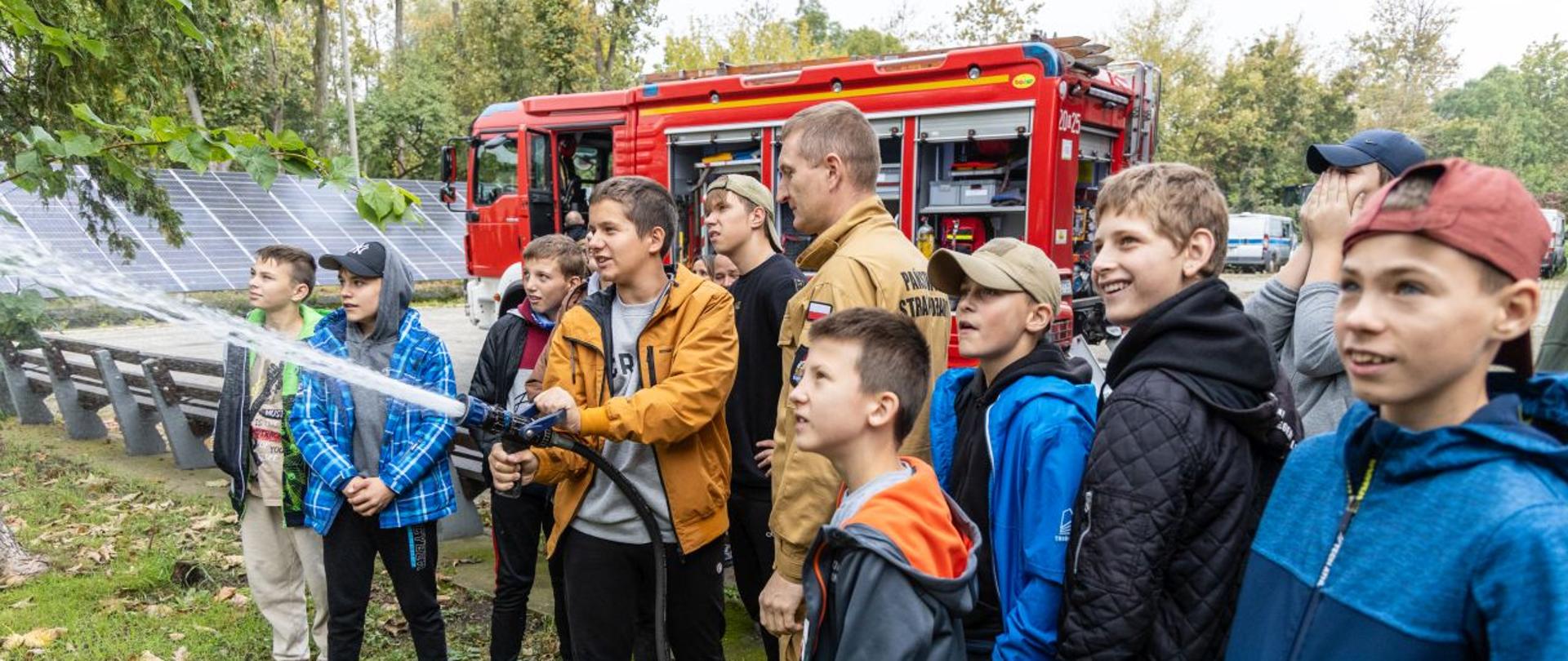 Strażak Komendy Wojewódzkiej PSP w Opolu pokazuje uczniom Młodzieżowego Ośrodka Socjoterapii w Dobrodzieniu działanie prądu gaśniczego z tzw. szybkiego natarcia. Uczniowie mogli spróbować poczuć się jak strażak i trzymając prądownice podać wodę na trawę. W tle widoczny samochód ratowniczo-gaśniczy Komendy Wojewódzkiej PSP w Opolu oraz instalację fotowoltaiczną.