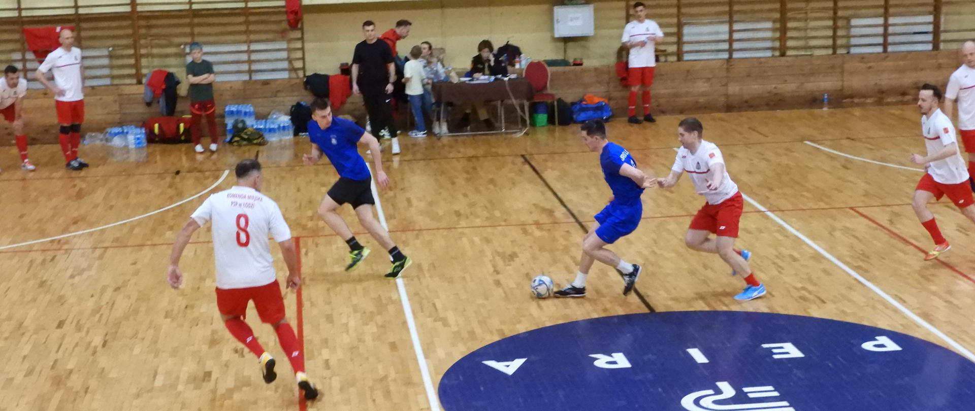 ecz piłki halowej rozgrywany w sali gimnastycznej. Kilku zawodników w dwóch drużynach – jedna w białych koszulkach i czerwonych spodenkach, druga w niebieskich strojach – walczy o piłkę na środku boiska. Jeden z graczy w niebieskim prowadzi piłkę, próbując minąć przeciwników. W tle widoczne są ławki, osoby siedzące przy stoliku oraz trybuny z kilkoma widzami
