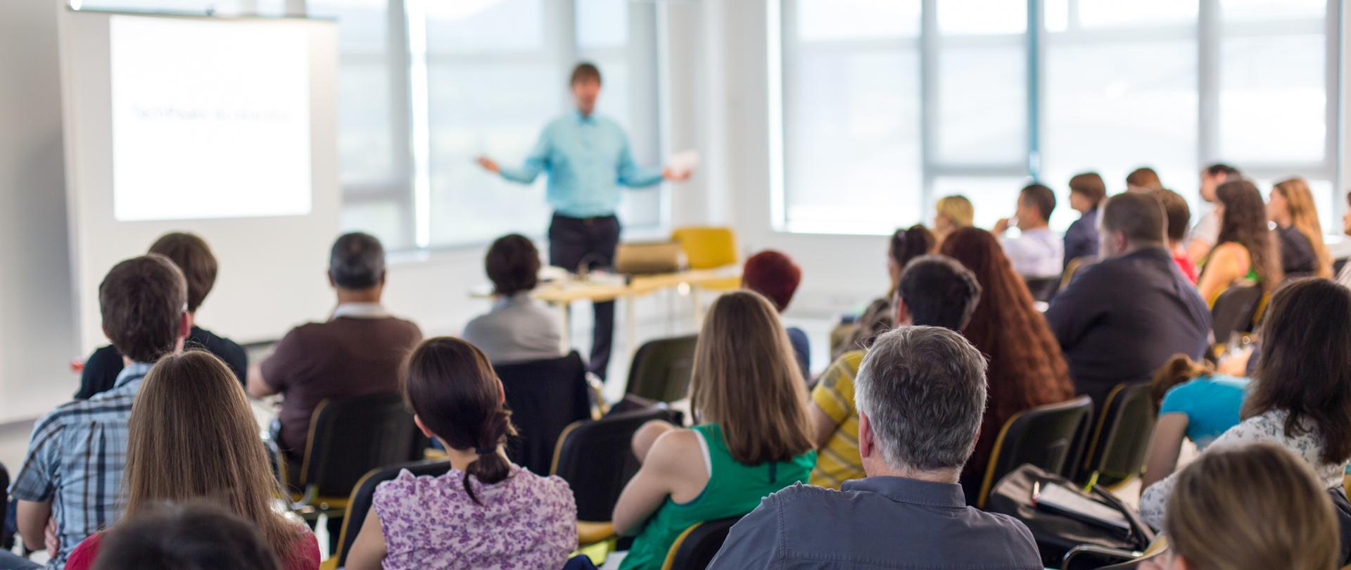 Business and entrepreneurship symposium. Speaker giving a talk at business meeting. Audience in conference hall. Rear view of unrecognized participant in audience.