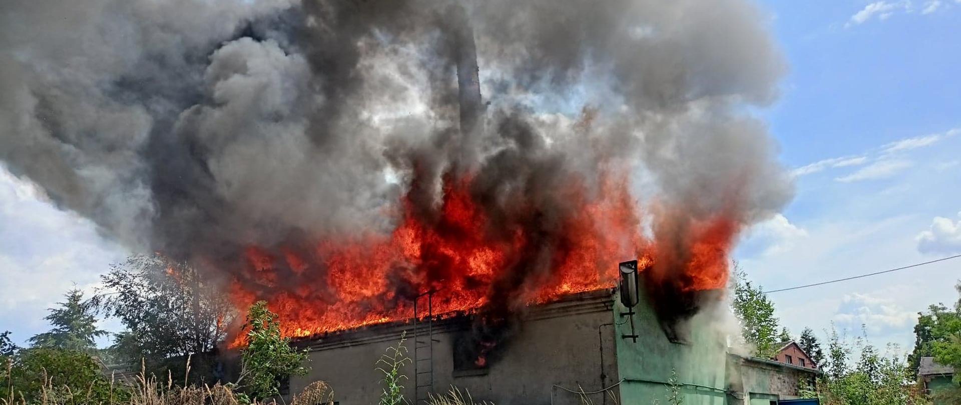 Zdjęcie przedstawia pożar budynku. Ogień wychodzi przez otwory okienne, pali się dach obiektu.