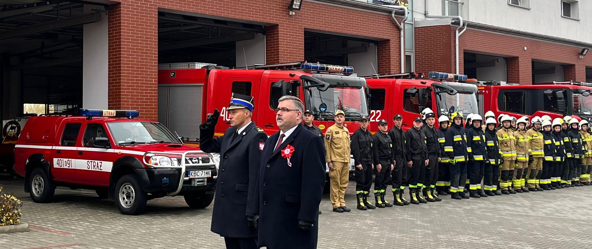 Zdjęcie przedstawia strażaków z Państwowej i Ochotniczej Straży Pożarnej w mundurach wyjściowych, strażaków ubranych ubrania koszarowe, koloru czarnego OSP. W tle znajdują się samochody pożarnicze koloru czerwonego, bramy garażowe i budynek strażnicy