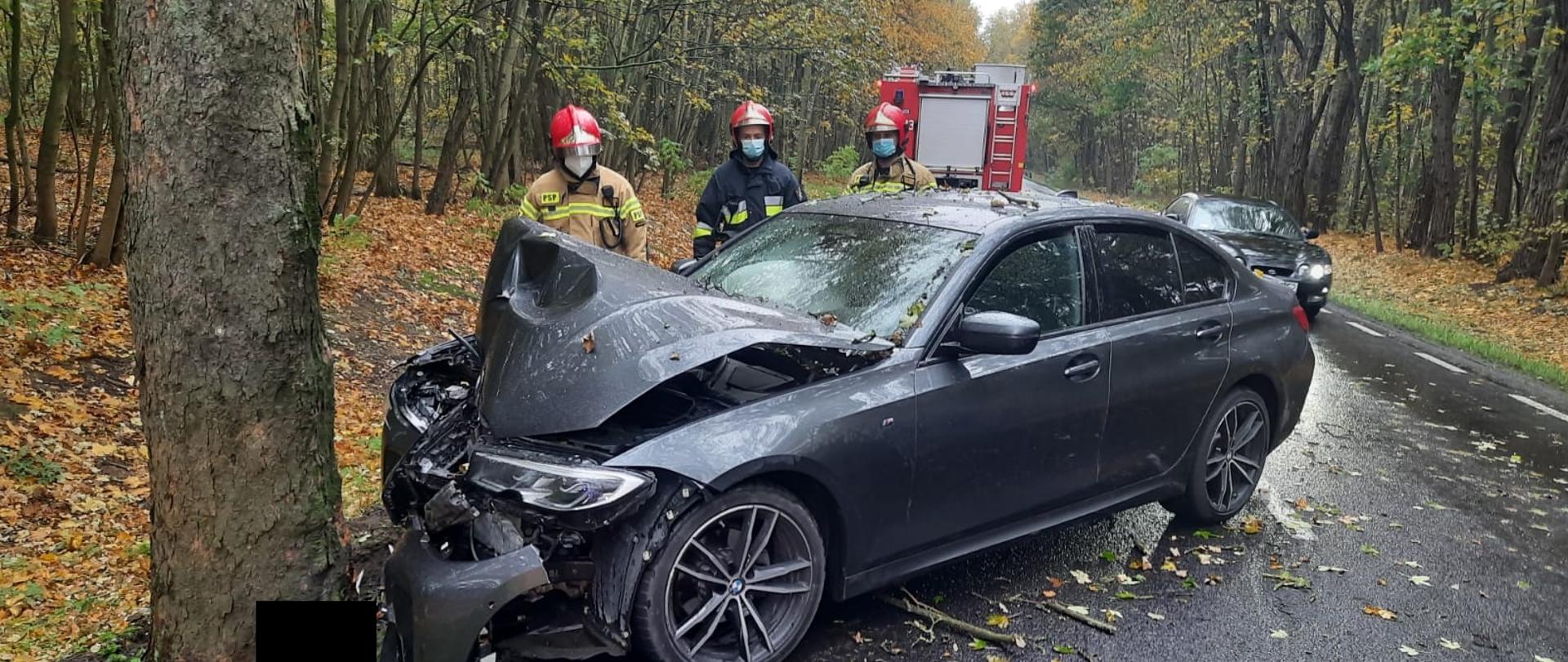 Ciemny samochód znajduje się przodem przy drzewie z uszkodzoną maską i zderzakiem, w pobliżu znajduje się trzech strażaków, w oddali stoi samochód straży pożarnej 