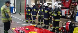 Widok z przodu. Siedmiu strażaków OSP w ubraniach specjalnych i hełmach stoi w szeregu przed matą z hydraulicznym sprzętem hydraulicznym. Przed strażakami stoi instruktor z PSP w ubraniu specjalnym. 