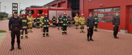 Zdjęcie strażaków w pozycji stojącej, na pierwszym planie trzech strażaków w umundurowaniu KDS, zaś na drugim planie 14 strażaków w umundurowaniu specjalnym. Wszyscy na planie budynku KP PSP w Brzezinach oraz samochodu bojowego z JRG Brzeziny 