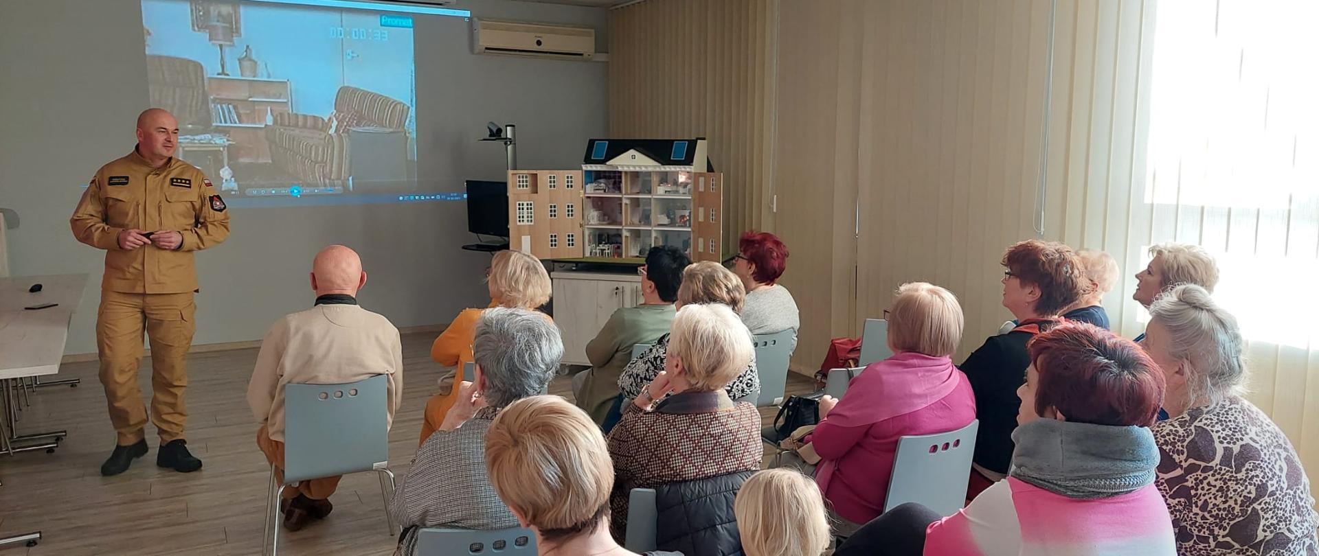 Zdjęcie przedstawia strażaka, który omawia seniorom zagrożenia występujące w budynkach mieszkalnych przy użyciu mobilnego symulatora zagrożeń pożarowych. Seniorzy siedzą na krzesełkach. 