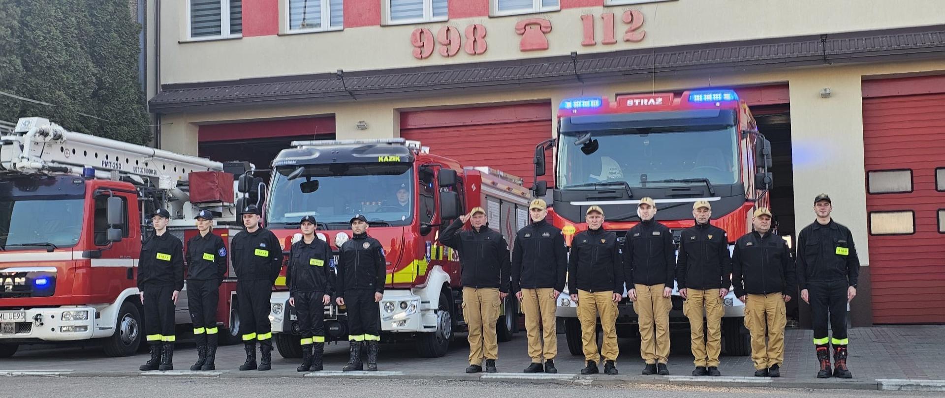 Strażacy ustawieni w szeregu przed budynkiem remizy strażackiej, na tle kilku czerwonych wozów bojowych z włączonymi sygnałami świetlnymi. Funkcjonariusze stoją na baczność, część w mundurach bojowych, część w ubraniach koszarowych. Nad garażami widoczny jest napis „998” oraz symbol telefonu. Zdjęcie przedstawia moment oddania hołdu – skupienie i powaga podkreślają uroczysty charakter chwili.
