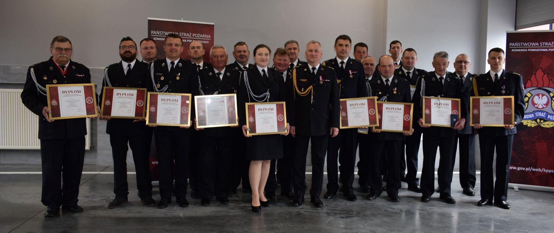 Powiatowe Obchody Dnia Strażaka w Łosicach. Zdjęcie grupowe strażaków w mundurach galowych stojących w pomieszczeniu garażowym. Strażacy trzymają w rękach dyplomy oprawione w złote cienkie ramki. 