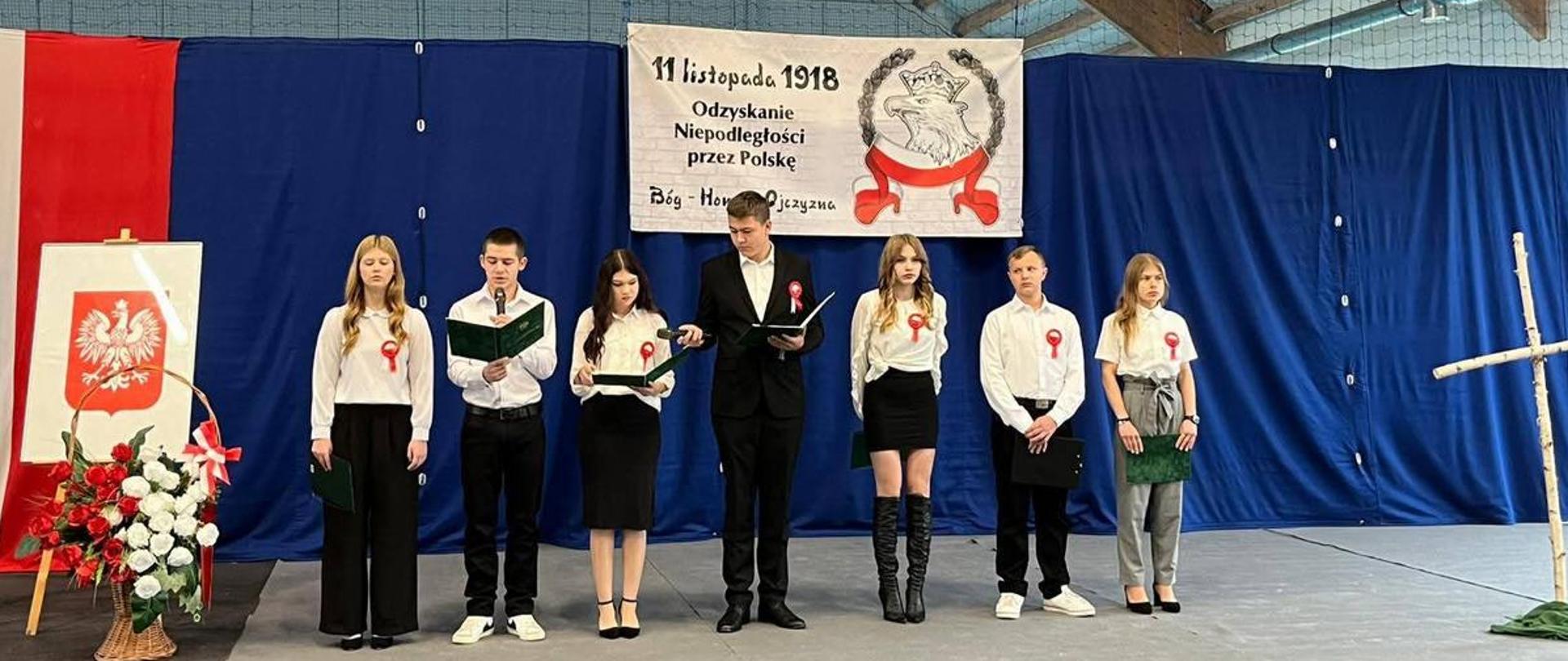 Siedmiu uczniów w strojach galowych (białe koszule, czarne spódnice/spodnie, kotyliony) stoi na tle niebieskiej kurtyny. Nad nimi wisi baner informujący: "11 listopada 1918 Odzyskanie Niepodległości przez Polskę". Dekoracje obejmują polską flagę, godło oraz brzozowy krzyż, podkreślając patriotyczny charakter uroczystości.