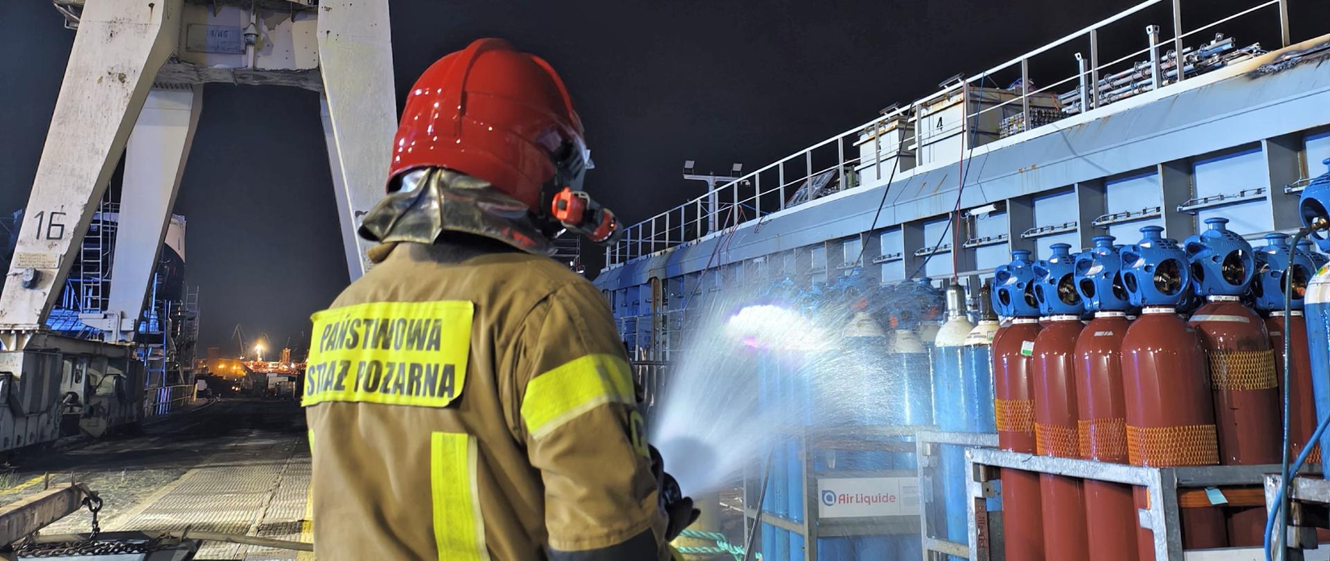 Strażak w ubraniu specjalnym PSP i czerwonym hełmie kieruje strumień wody w stronę ustawionych pionowo butli gazowych (Air Liquide) na pokładzie jednostki. Akcja odbywa się nocą w rejonie portowym, a w tle widoczny jest wysoki żuraw stoczniowy.