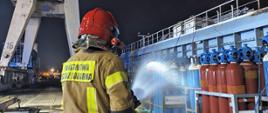 Strażak w ubraniu specjalnym PSP i czerwonym hełmie kieruje strumień wody w stronę ustawionych pionowo butli gazowych (Air Liquide) na pokładzie jednostki. Akcja odbywa się nocą w rejonie portowym, a w tle widoczny jest wysoki żuraw stoczniowy.