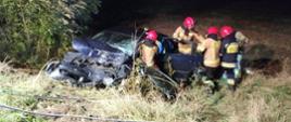 Zdjęcie zrobione w nocy. Rozbite auto koloru granatowego stoi w rowie skierowane uszkodzonym przewodem w aparat. Miejsce oświetlone lampą. Przy aucie stoi 5 strażaków w umundurowaniu bojowym, strażacy wykonują dostęp do zakleszczonego kierowcy