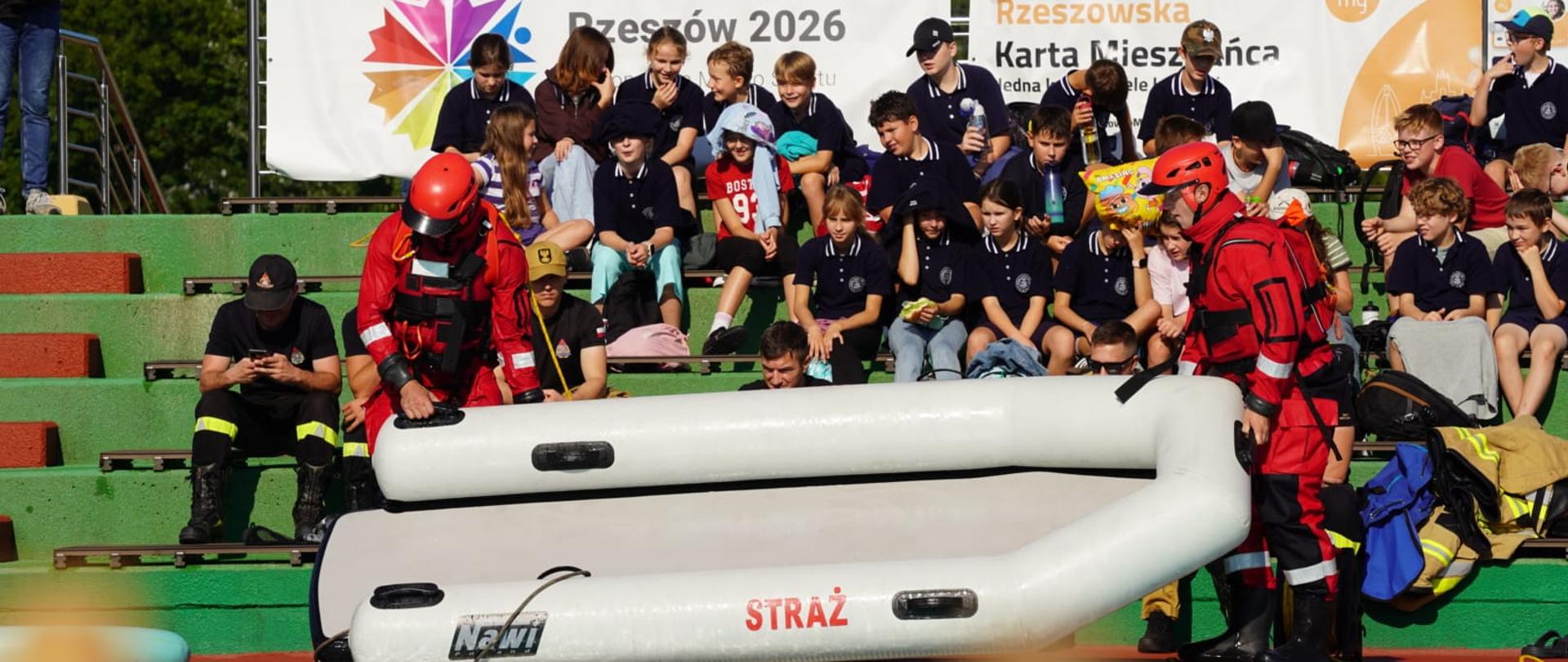 Dwóch ratowników w czerwonych kombinezonach przygotowuje ponton z napisem „STRAŻ” do pokazowego wodowania przy brzegu basenu. W tle, na trybunach, siedzi grupa uczniów ubranych w granatowe koszulki z logo szkoły, obserwująca przebieg ćwiczeń. Za nimi widnieje baner z napisem „Rzeszów 2026” oraz „Rzeszowska Karta Mieszkańca”. Wydarzenie ma charakter edukacyjny i promuje zasady bezpieczeństwa nad wodą oraz pracę służb ratowniczych.