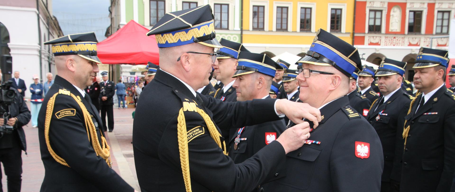 Wyróżnieni strażacy stojący w szyku. Na pierwszym planie zastępca komendanta głównego PSP wręczający odznaczenie.