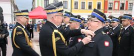Wyróżnieni strażacy stojący w szyku. Na pierwszym planie zastępca komendanta głównego PSP wręczający odznaczenie.