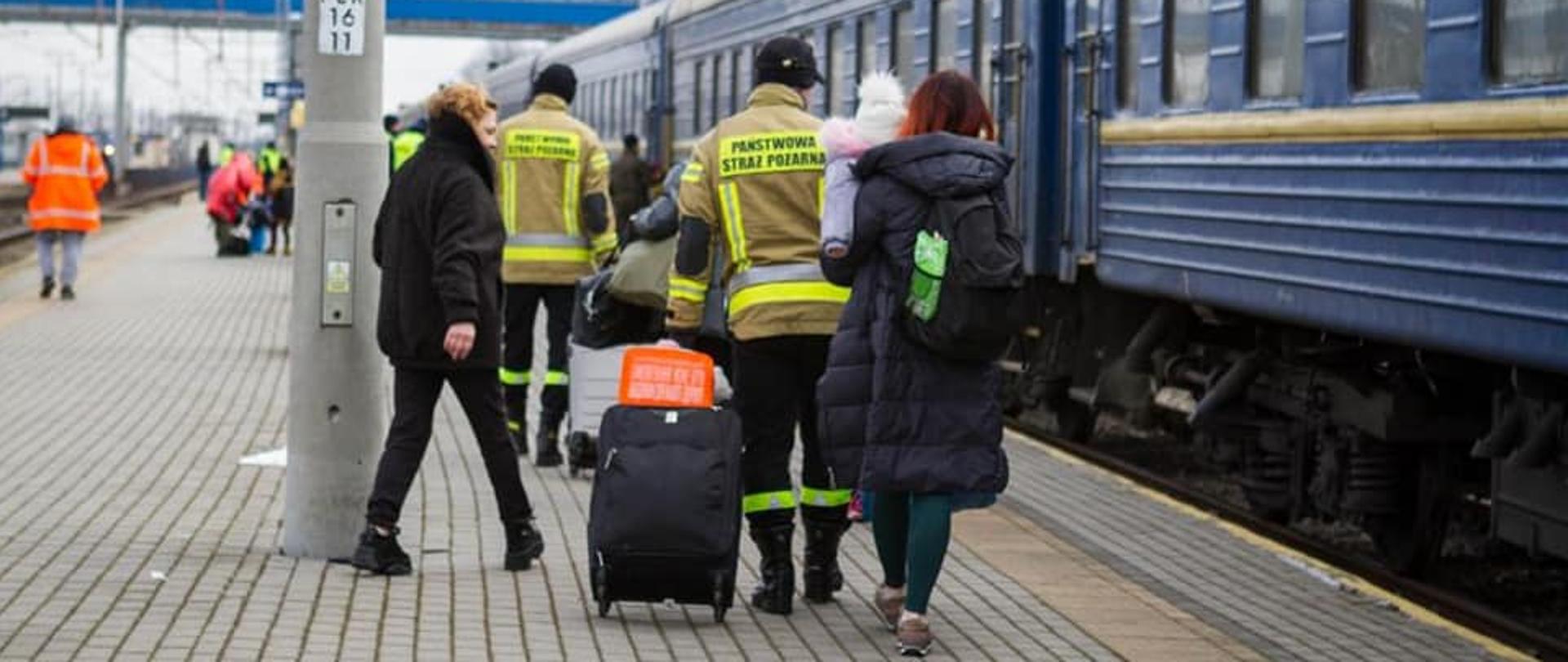 Na zdjęciu widać 2 strażaków PSP na peronie PKP pomagających w przemieszczaniu się osobom z Ukrainy. Strażacy prowadzą walizki. 