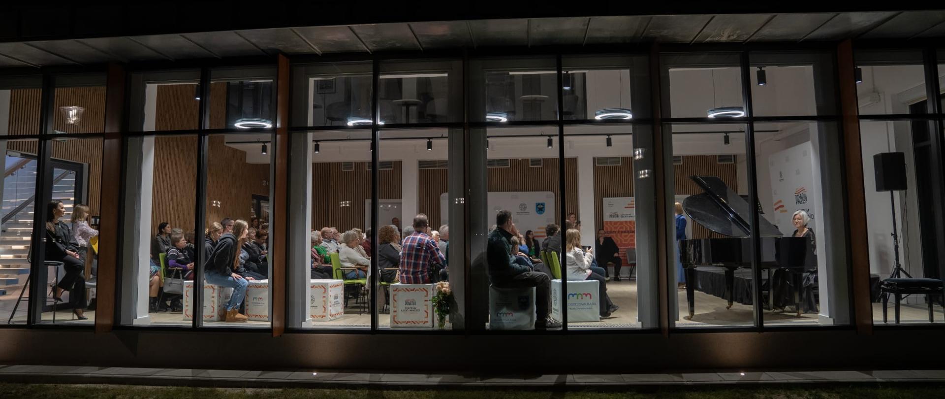 Na zdjęciu widać nowoczesny budynek z dużymi przeszklonymi oknami, przez które można
zobaczyć wnętrze oświetlonego pomieszczenia. W środku znajduje się publiczność siedząca
na krzesłach, zwrócona w stronę sceny, na której stoi fortepian. Przy fortepianie siedzi
kobieta, prawdopodobnie pianistka, gotowa do występu lub już grająca. Pomieszczenie jest
elegancko urządzone, z nowoczesnym oświetleniem sufitowym i drewnianymi elementami
dekoracyjnymi na ścianach.
Zdjęcie zostało wykonane z zewnątrz, w nocy, co podkreśla kontrast między ciemnością na
zewnątrz a jasnym wnętrzem budynku. Możliwe, że jest to koncert, recital lub inne
wydarzenie kulturalne.
