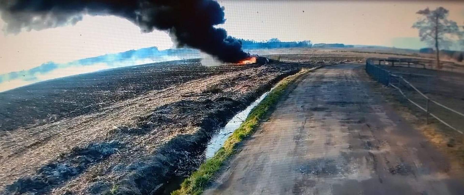 Na zdjęciu pole oraz polna droga. Na środku stos palących się opon. Duży, czarny dym. Zachmurzenie duże. W tle drzewa.