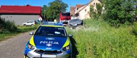 Radiowóz Policji stojący na drodze. W tle widoczny jest uszkodzony samochód osobowy oraz wóz strażacki.