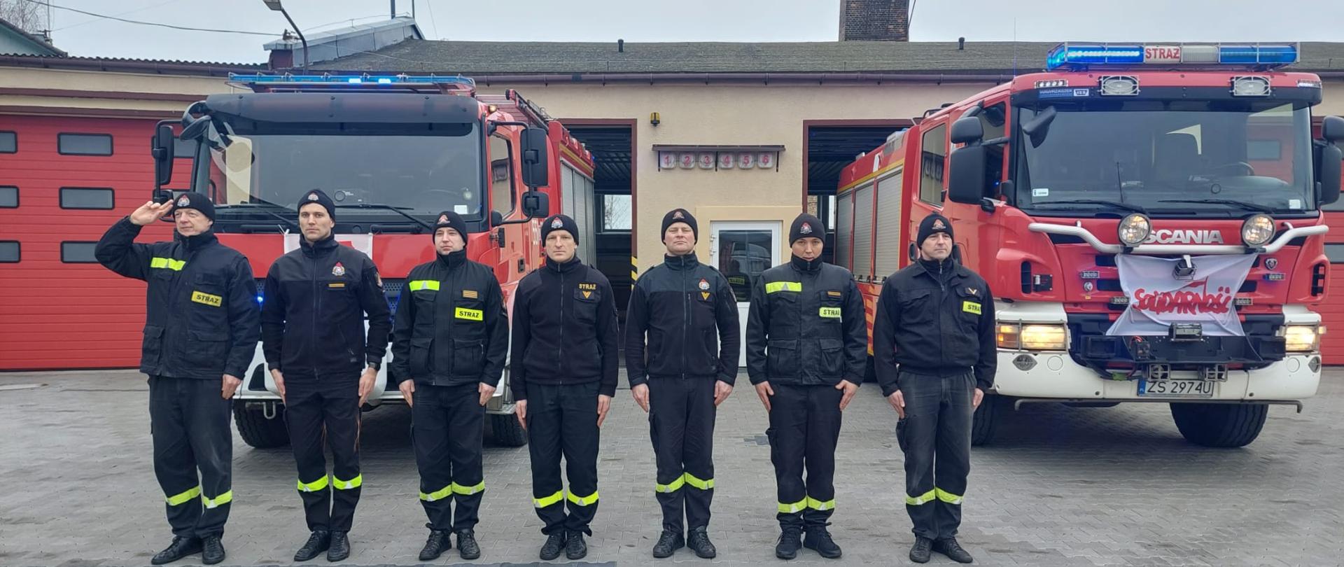 Zdjęcie przedstawia dwa wozy strażackie przed bramami garażowymi. Przed samochodami stoją strażacy na baczność oddając hołd zmarłemu strażakowi.