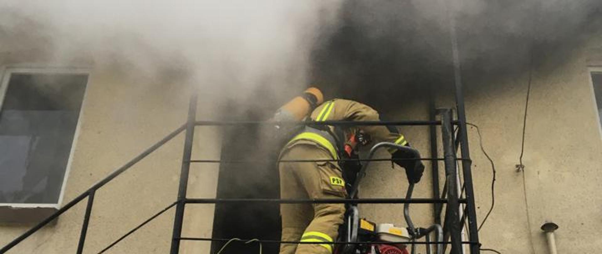 Strażak zabezpieczony aparatem ochrony dróg oddechowych stoi na spoczniku zewnętrznych metalowych schodów i uruchamia wentylatro osiowy, aby oddymić pomieszczenia