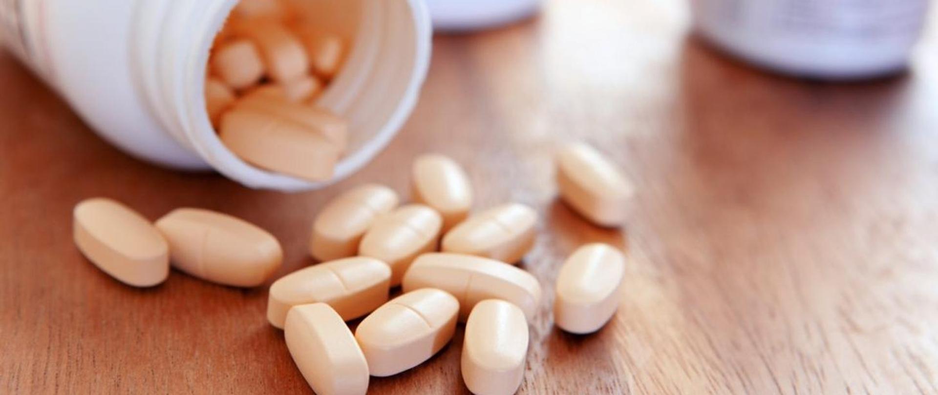 Na zdjęciu widoczna jest plastikowa butelka z suplementami diety lub lekami, leżąca na boku na drewnianej powierzchni. Z butelki wysypały się beżowe tabletki o podłużnym kształcie, z wyraźnym rowkiem dzielącym każdą z nich na pół. W tle, lekko rozmytym, widać jeszcze jedną podobną butelkę. 