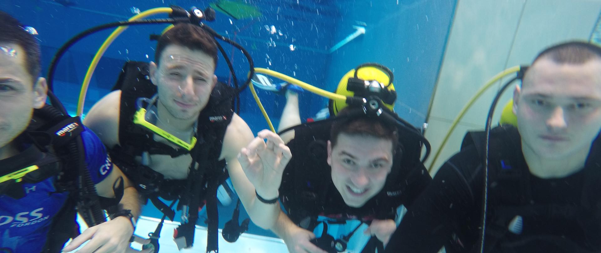 Czterech nurków pod wodą na bezdechu, na plecach butle nurkowe. Wszyscy twarzami skierowani do osoby robiącej zdjęcie.