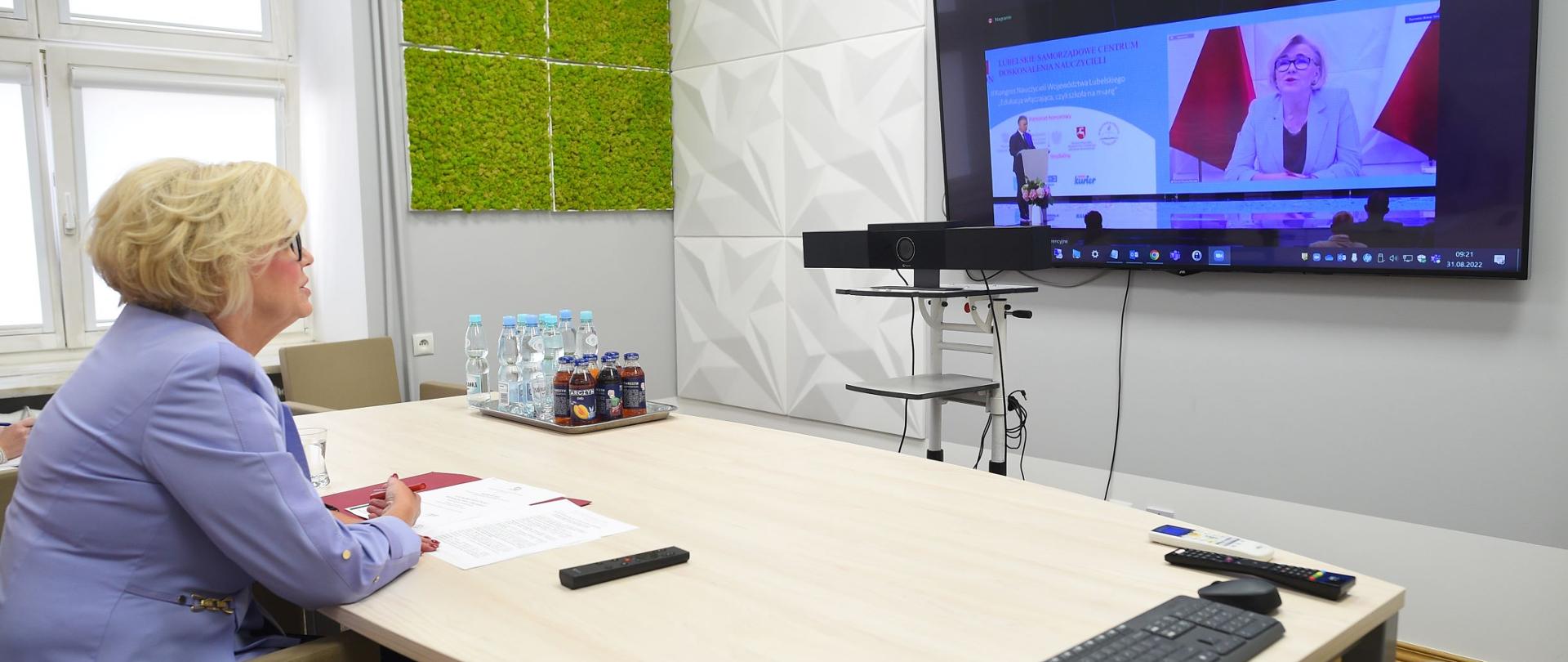 Wiceminister Marzena Machałek siedzi przy stole, przed ekranem telewizora na którym transmitowana jest konferencja.