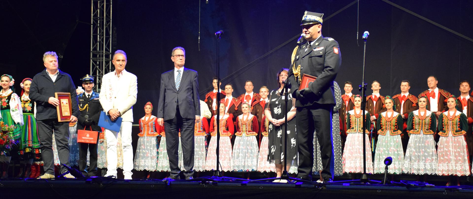 Na zdjęciu grupa osób stojąca na scenie. Po prawej stronie, w mundurze galowym do mikrofonu przemawia Śląski Komendant Wojewódzki Państwowej Straży pożarnej nadbrygadier Jacek Kleszczewski. W tle kobiety z Zespołu Pieśni i Tańca Śląsk ubrane w stroje ludowe.