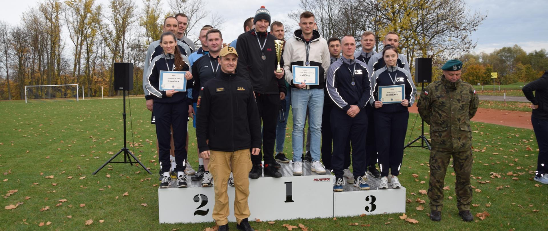 Na pierwszym planie zdjęcia widzimy przedstawicieli służb mundurowych stojących na podium po zakończonych zawodach sportowych. Na drugim planie widzimy boisko sportowe. 