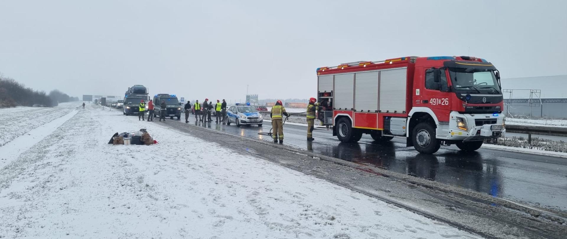 Na zdjęciu znajduje się wrak spalonego pojazdu, strażacy w ubraniach specjalnych, wozy strażackie oraz radiowóz