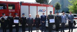 Strażacy oraz goście dokonujący przekazania pojazdów pożarniczych (pojazdy w tle)