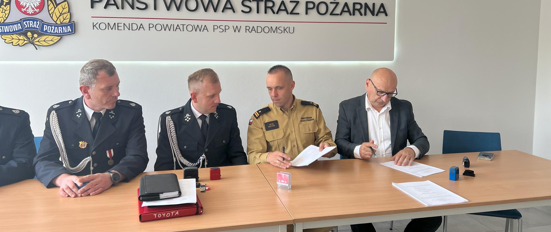 Na zdjęciu 4 osoby siedzące za biurkiem podczas podpisywaniu dokumentów w tle logo PSP oraz napis PAŃSTWOWA STRAŻ POŻARNA a także godło Polski 