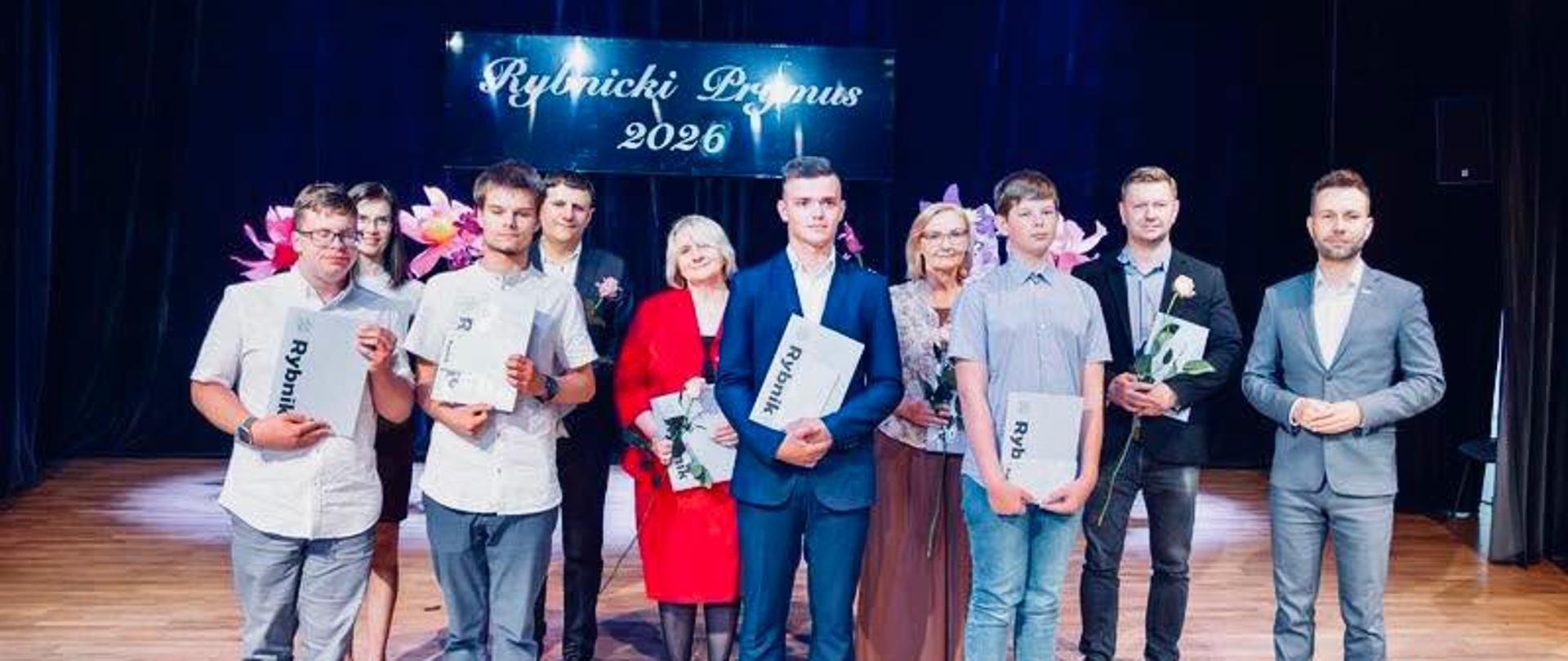 Grupowe zdjęcie Rybnickich Prymusów 2026 na scenie. Dziesięć osób - 4 uczniów oraz ich nauczyciele i Wiceprezydent miasta Rybnika - stoi w dwóch rzędzie na drewnianej scenie z ciemną kurtyną w tle. W centrum widoczny jest napis świetlny "Rybnicki Prymus 2026". Prymusi, młodzi mężczyźni w casualowych i eleganckich strojach, trzymają dyplomy i nagrody. Wśród nich znajdują dorośli - trzy kobiety w eleganckich strojach (garsonkach) oraz trzech mężczyzn w garniturach.