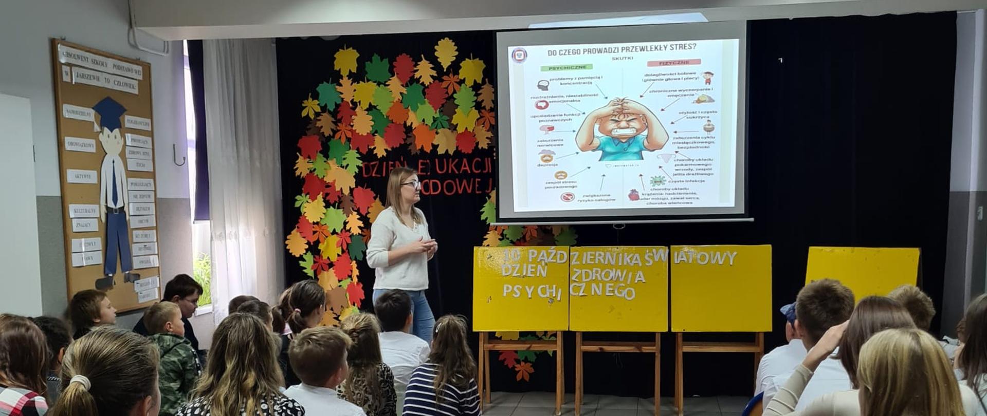 . W centralnej części zdjęcia widoczny jest ekran, na którym wyświetlana jest prezentacja. Pod ekranem znajdują się 4 żółte tablice, na których wcześniej uczniowie ułożyli biały napis „10 października Światowy Dzień Zdrowia Psychicznego”, po lewej stronie tablic stoi pracownica PSSE w Kamieniu Pomorskim prowadząca prelekcję. Do ekranu prowadzi wąski czerwony dywan a po jego obu stronach na krzesłach siedzą uczniowie Szkoły Podstawowej w Jarszewie.