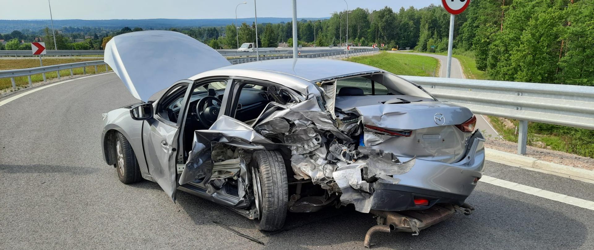 Zdjęcie obrazuje uszkodzony samochód osobowy Mazda. Siła z jaką uderzył w niego samochód Nissan była tak duża, że tylne koło przemieściło się aż do foteli tylnej kanapy pasażerskiej. Cała karoseria lewego tyłu pojazdu jest sprasowana. Szyby w pojeździe są powybijane. Przednia maska jest podniesiona do góry. W tle widać panoramę w oddali las, a w bliższej perspektywie infrastrukturę drogową.