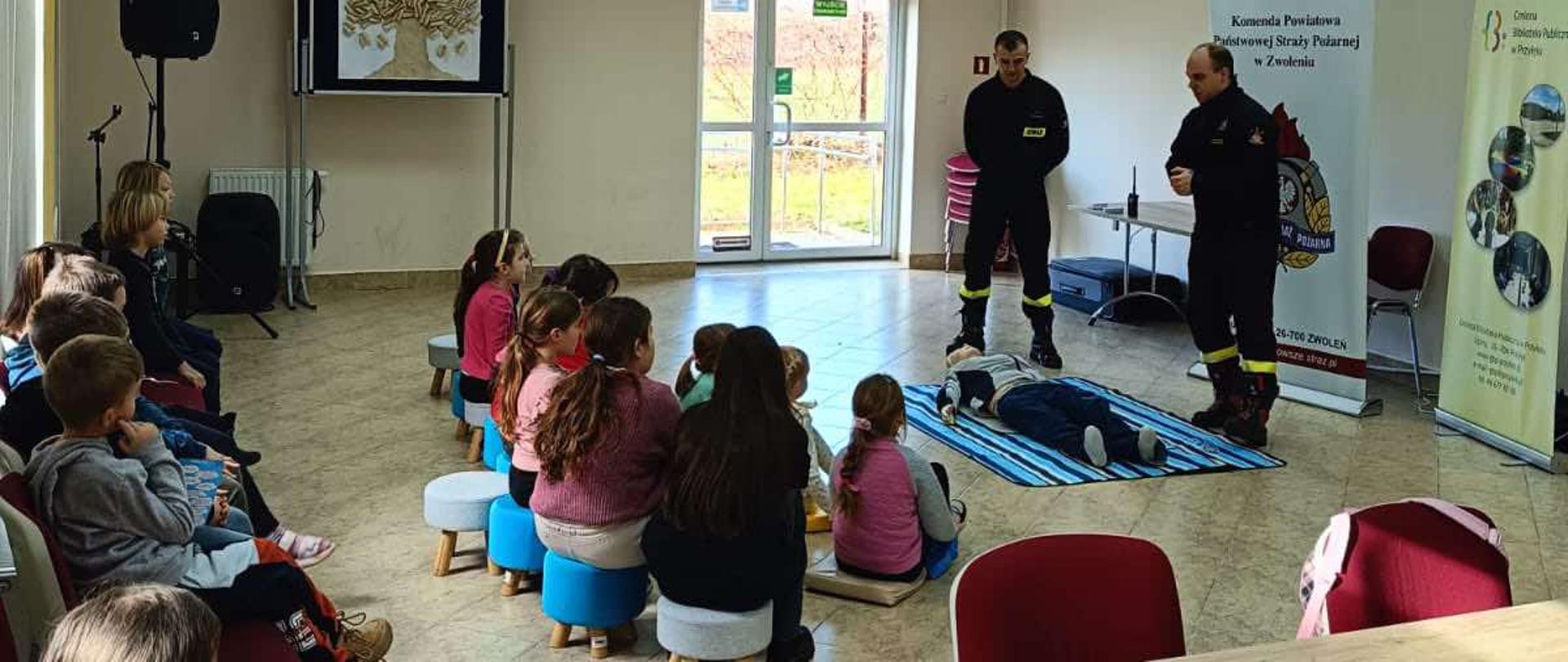 Na zdjęciu widoczny jest pokaz pierwszej pomocy prowadzony przez dwóch strażaków dla grupy dzieci w pomieszczeniu, prawdopodobnie sali szkolnej lub świetlicy. Jeden ze strażaków klęczy obok manekina leżącego na niebieskiej macie, demonstrując techniki pierwszej pomocy, podczas gdy drugi stoi obok z mikrofonem, prawdopodobnie objaśniając czynności. Dzieci siedzą na krzesłach i małych taboretach, uważnie obserwując pokaz. W tle widoczny jest baner z napisem "Komenda Powiatowa Państwowej Straży Pożarnej w Zwoleniu" oraz informacjami kontaktowymi i logo PSP.