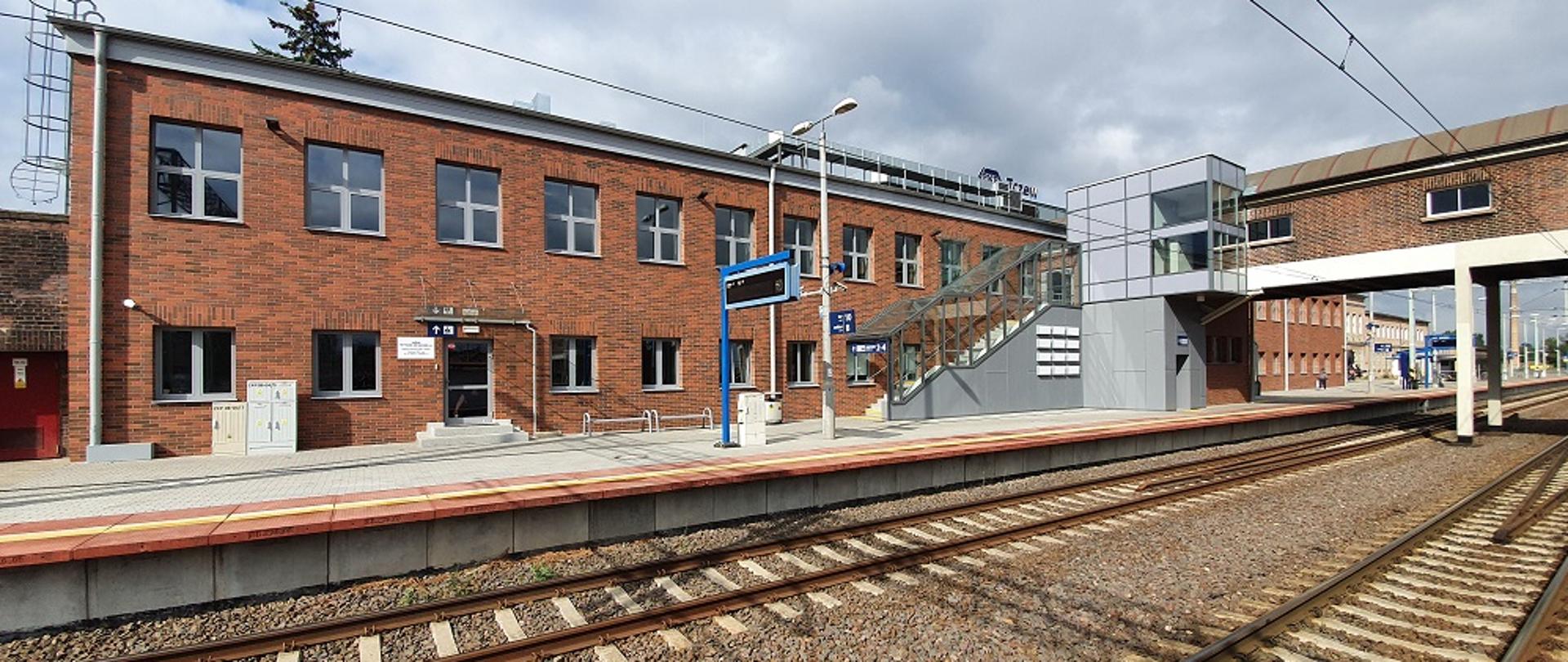 Na zdjęciu widoczny budynek dworca z ceglastą elewacją, peron oraz dwa tory, schody i windę.