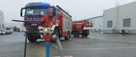 Stojak hydrantowy wraz z wężem podczas działań gaśniczych. W tle dwa pojazdy pożarnicze i hala produkcyjna w zakładzie.