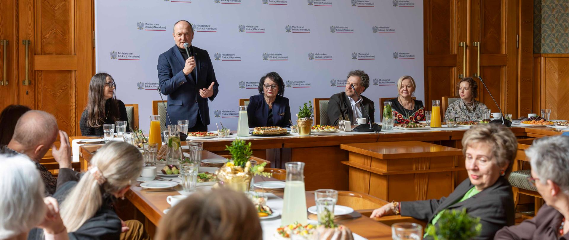 Mężczyzna w ciemnej marynarce i czarnym golfie przemawia do mikrofonu, stojąc za stołem. Po jego bokach siedzą inni uczestnicy spotkania. Na stole widoczne są tradycyjne potrawy wielkanocne, w tym faszerowane jajka i ciasta. W tle biała ścianka z powtarzającym się logo Ministerstwa Edukacji Narodowej.
