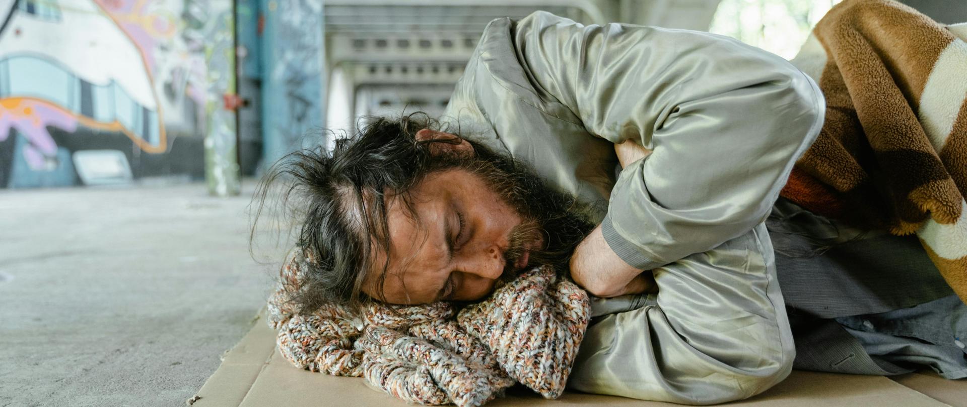 grafika przedstawia mężczyznę w kryzysie bezdomności leżącego pod kocem na chodniku