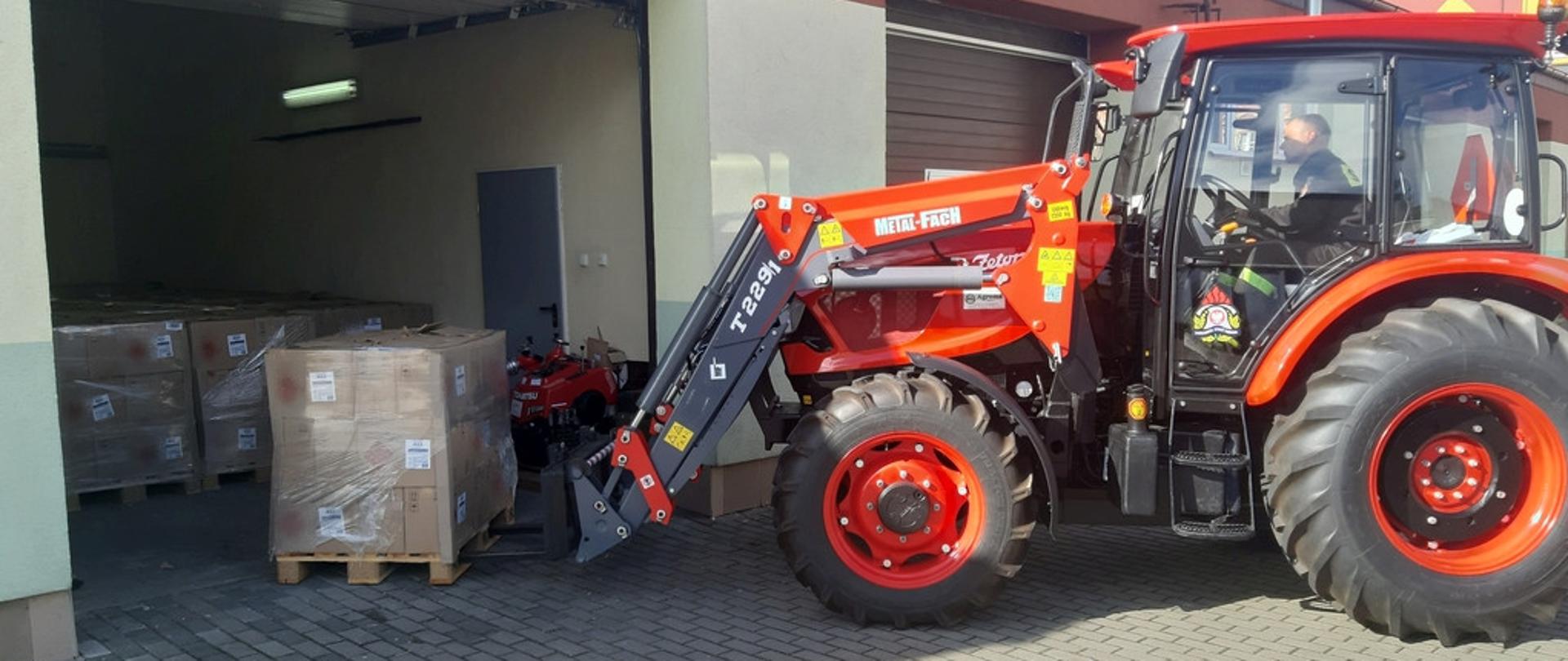 Na zdjęciu strażak za pomocą traktora z podnośnikiem przeładowuje środki ochrony indywidualnej zapakowane w kartony, na palecie