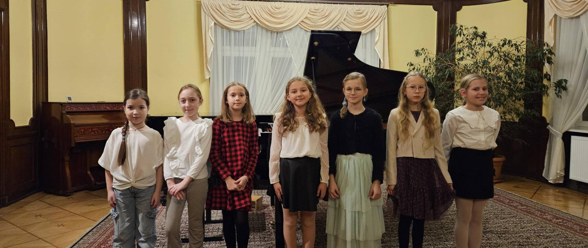 Zdjęcie kolorowe, sześć dziewcząt stojących obok siebie w rzędzie w sali, przed koncertem lub występem, w tle czarny fortepian, duża roślina doniczkowa i okna zasłonięte beżowymi, ozdobnymi zasłonami, na podłodze ozdobny dywan, sala z jasnymi, drewnianymi panelami na ścianach.