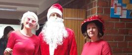 Mężczyzna stoi w korytarzu szkoły w stroju Mikołaja, po obu jego stronach stoją dwie kobiety w czerwonych elfów.