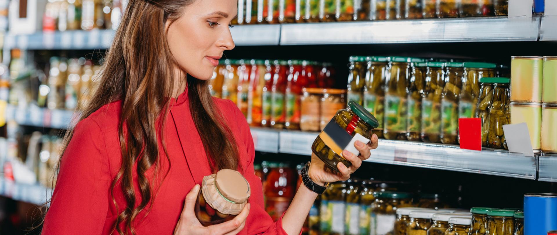 Na zdjęciu jest kobieta w sklepie, która trzyma w dłoniach dwa słoiki. Spogląda na jednego z nich.