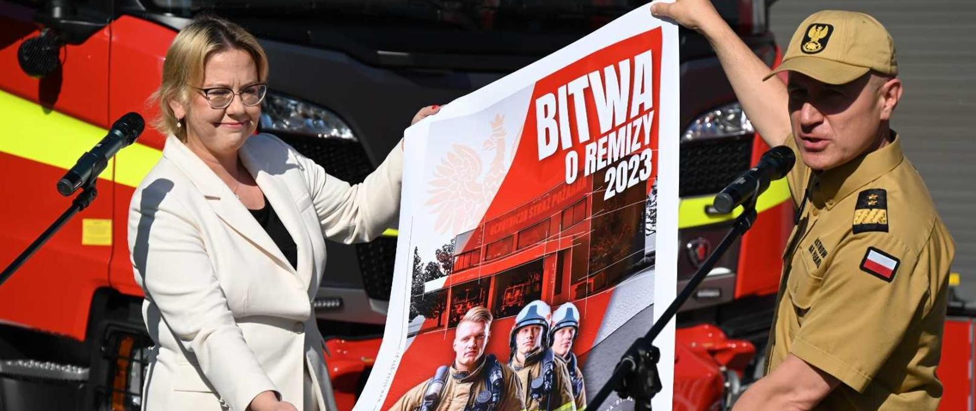 Zdjęcie przedstawią Panią Minister w białej marynarce trzymającą plakat wraz z Komendantem Głównym Straży Pożarnej. Na plakacie strażacy i napis Bitwa o remizę 2023.