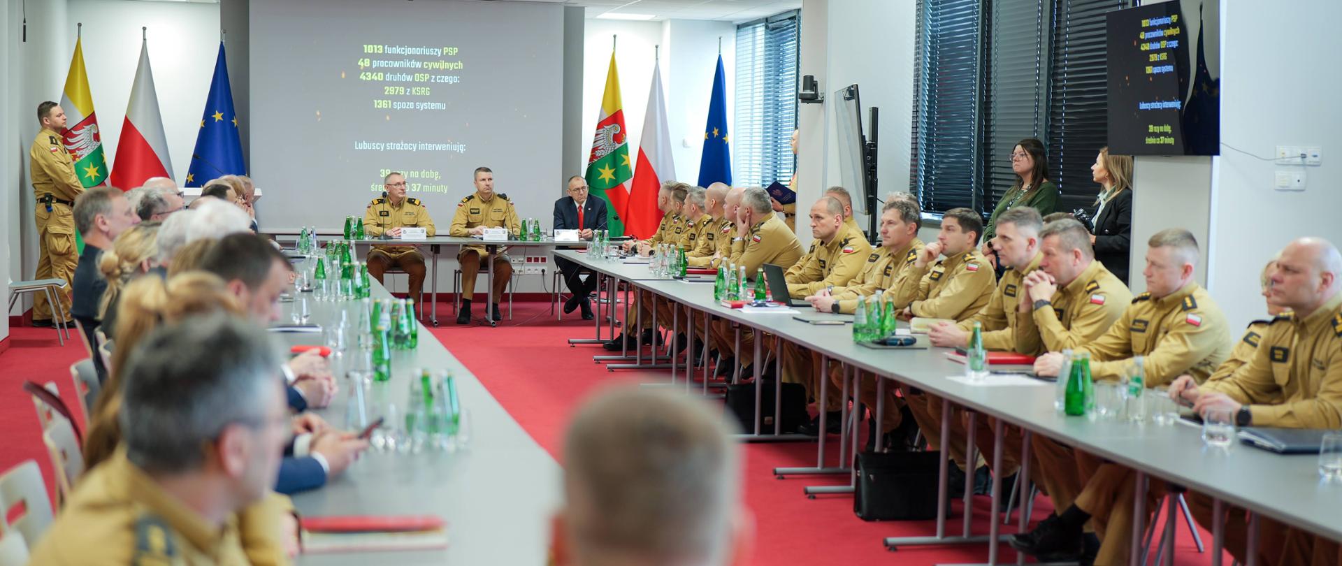 Szerokie ujęcie sali podczas odprawy rocznej. Funkcjonariusze PSP siedzą przy stołach ustawionych w podkowę, na ekranie wyświetlane są dane statystyczne dotyczące działalności straży pożarnej w regionie.
