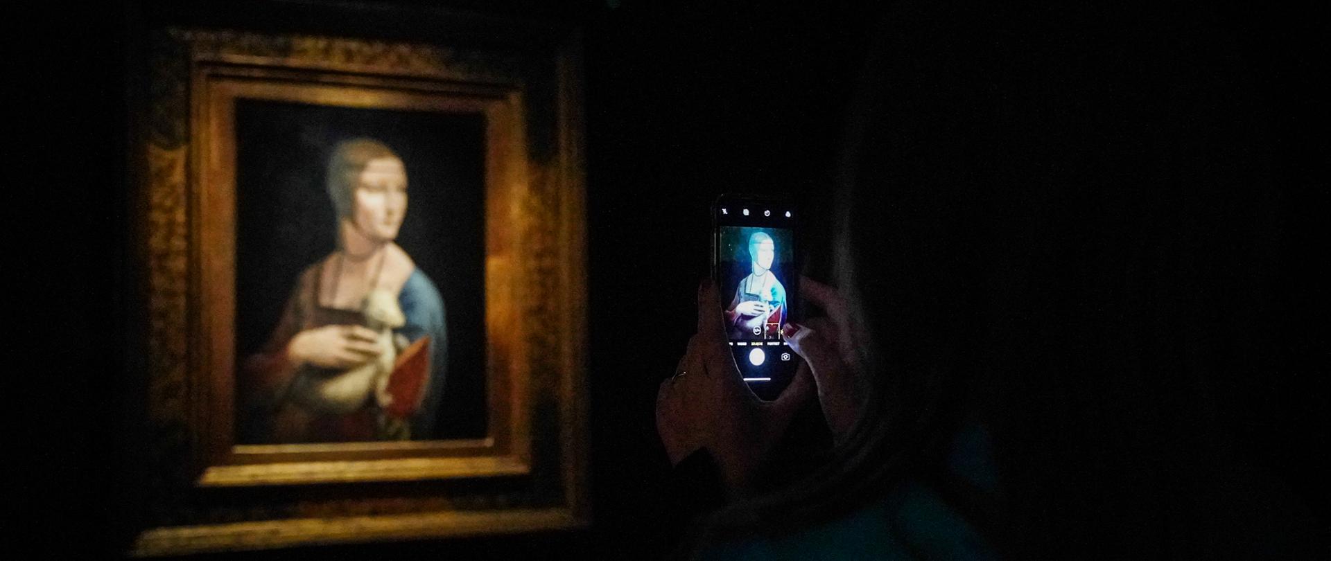 w zacienionej sali ekspozycyjnej ręce trzymają telefon komórkowy, którym robione jest zdjęcie widocznego obrazu "Damy z gronostajem" Leonarda Vinci; na obrazie kobieta pozuje z leżącym na ręce zwierzęciem, które przytrzymuje drugą ręką